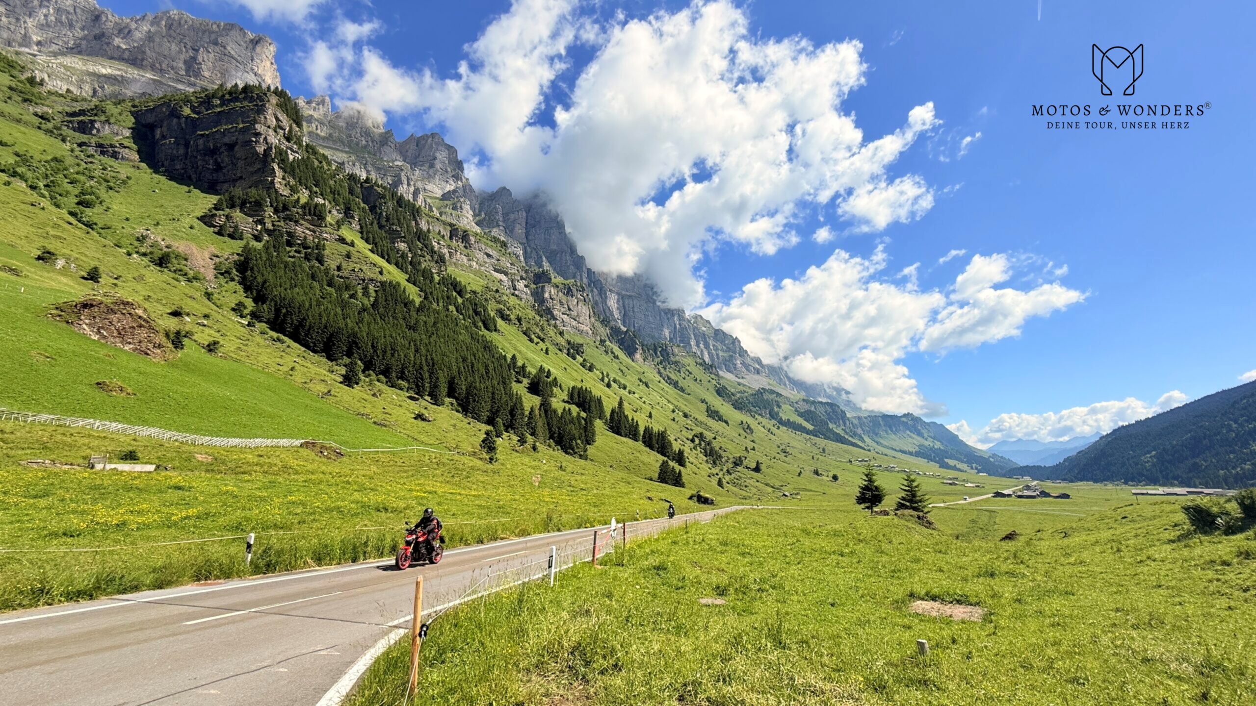 Motorradfahrer auf einer Strasse durch ein grünes Alpental mit steilen Felswänden und blauem Himmel.