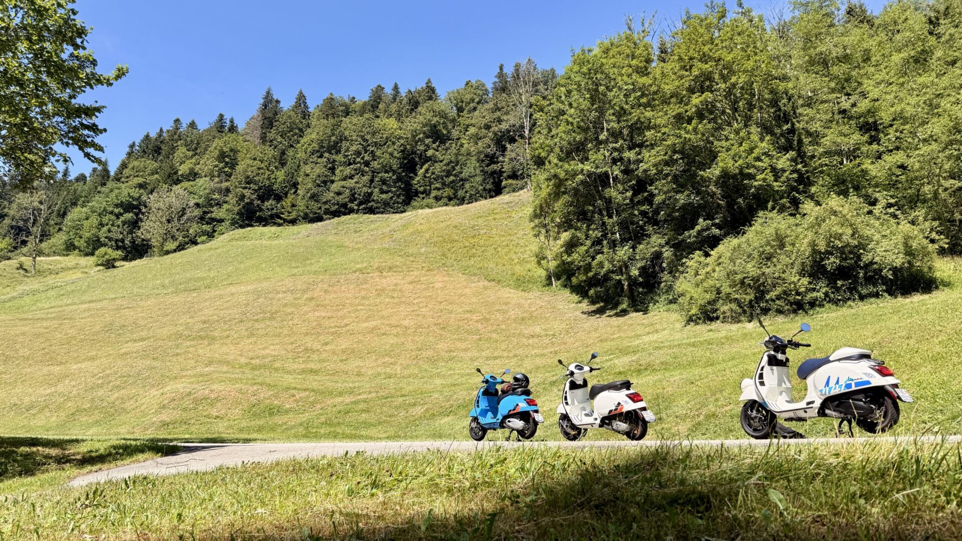 Drei Vespa-Roller am Wegrand vor grüner Hügelwiese und Wald im Emmental
